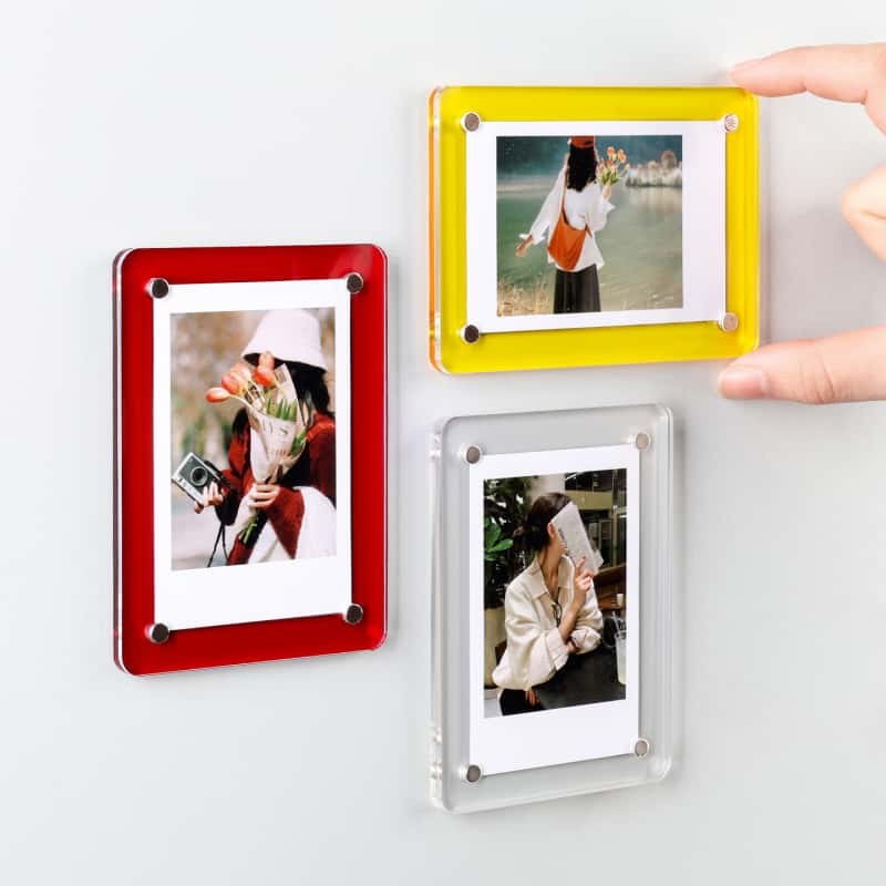 Red, yellow and clear acrylic magnetic picture frames on a steel refrigerator door, with a hand lifting one frame off the surface