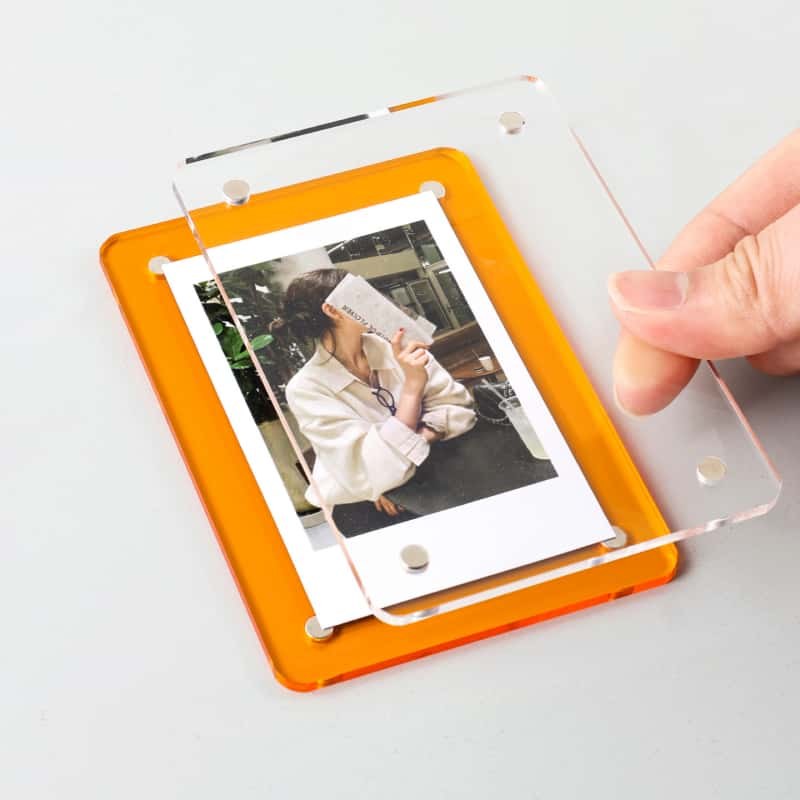 An acrylic magnetic picture frame opened in the hand, showing the front sheet, the orange rear sheet and the corner magnets that clamp the print
