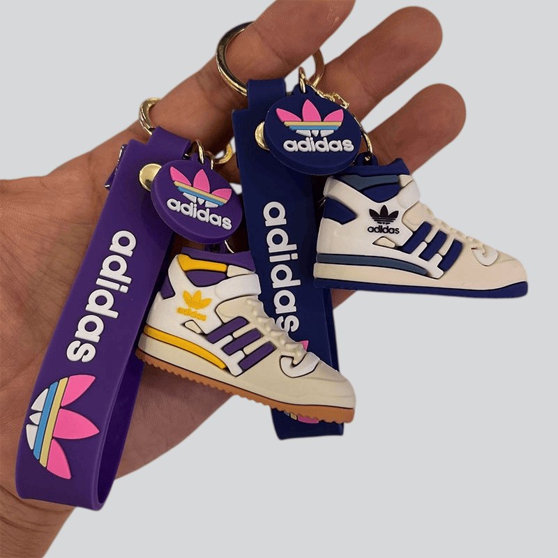 Adidas shoe keychains in purple and navy held in one hand, showing the tag strap printed with lettering