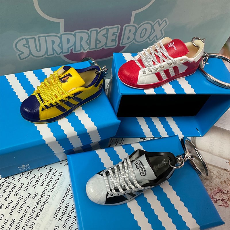 Three basketball shoe keychains in yellow, red and black resting on printed gift boxes in front of a display backdrop