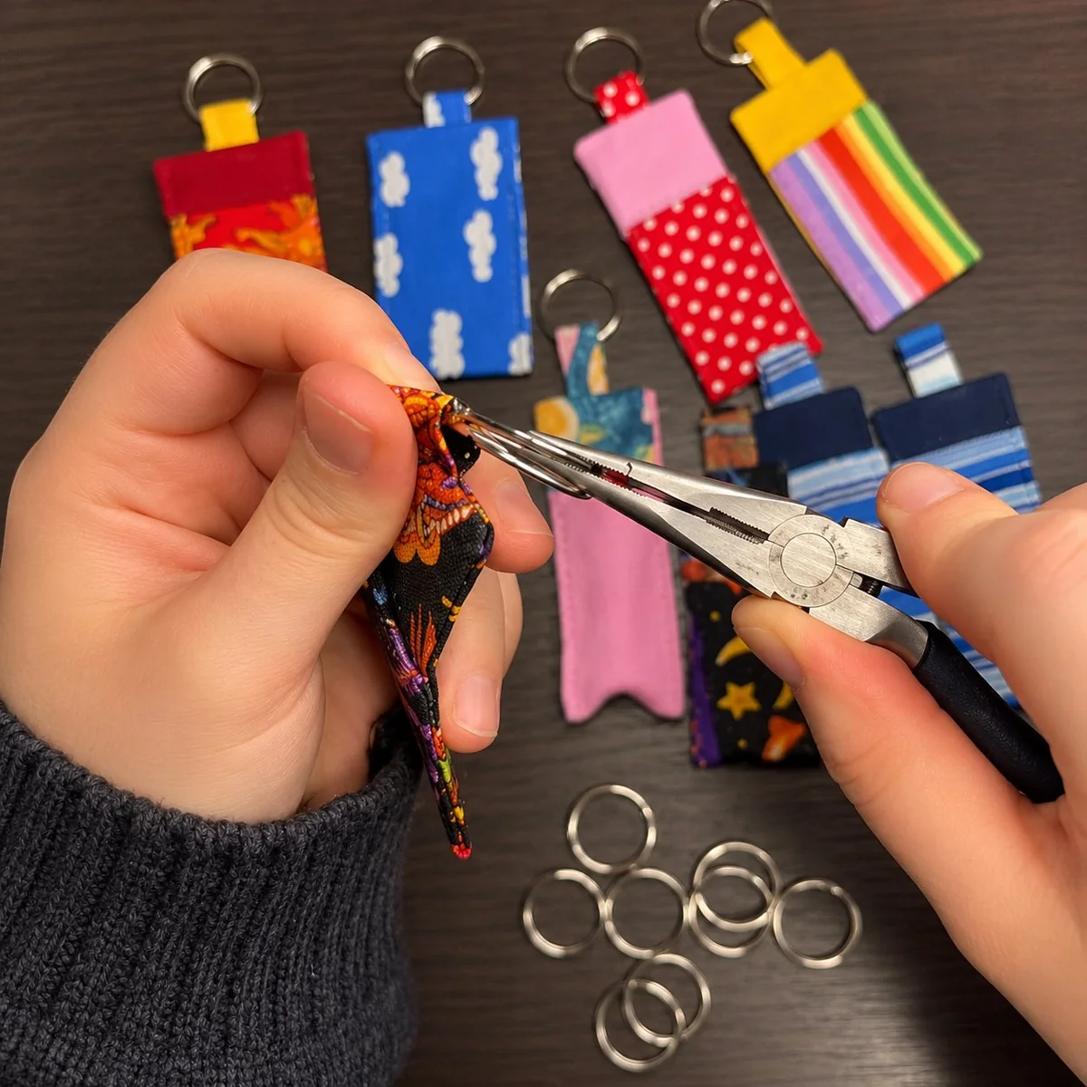 Hand holding flat-nose pliers and sliding a split ring onto a fabric keychain strap loop — using a tool on both sides keeps the ring round and avoids bending the metal.