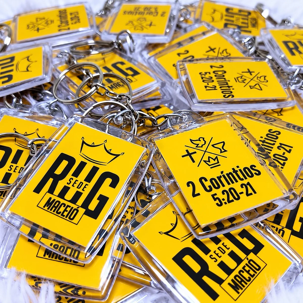 Yellow acrylic church event keychains — pile of clear acrylic tags printed with a '2 Coríntios 5:20-21' design
