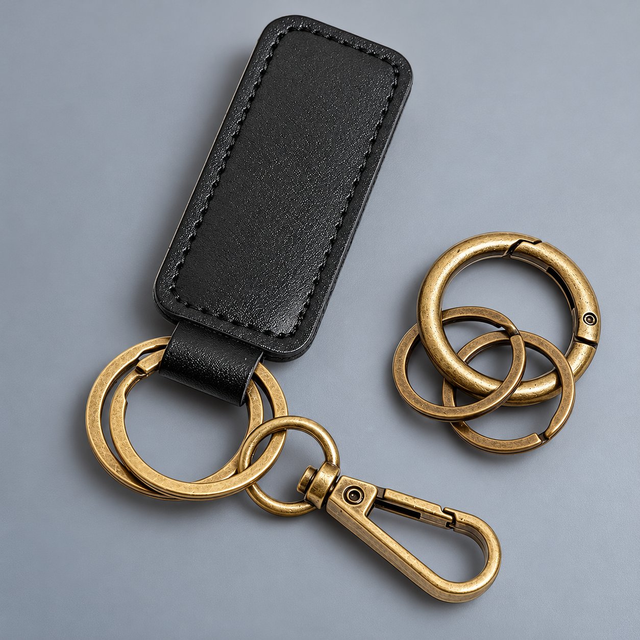 A black leather keychain strap with a stitched edge, brass split rings and a brass swivel snap hook on a grey background