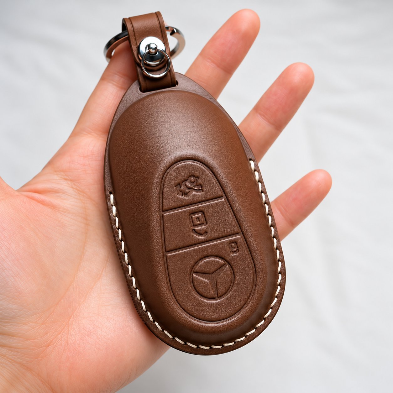 A hand holding a brown leather car key fob case with white stitching and the Mercedes-Benz emblem debossed into the surface