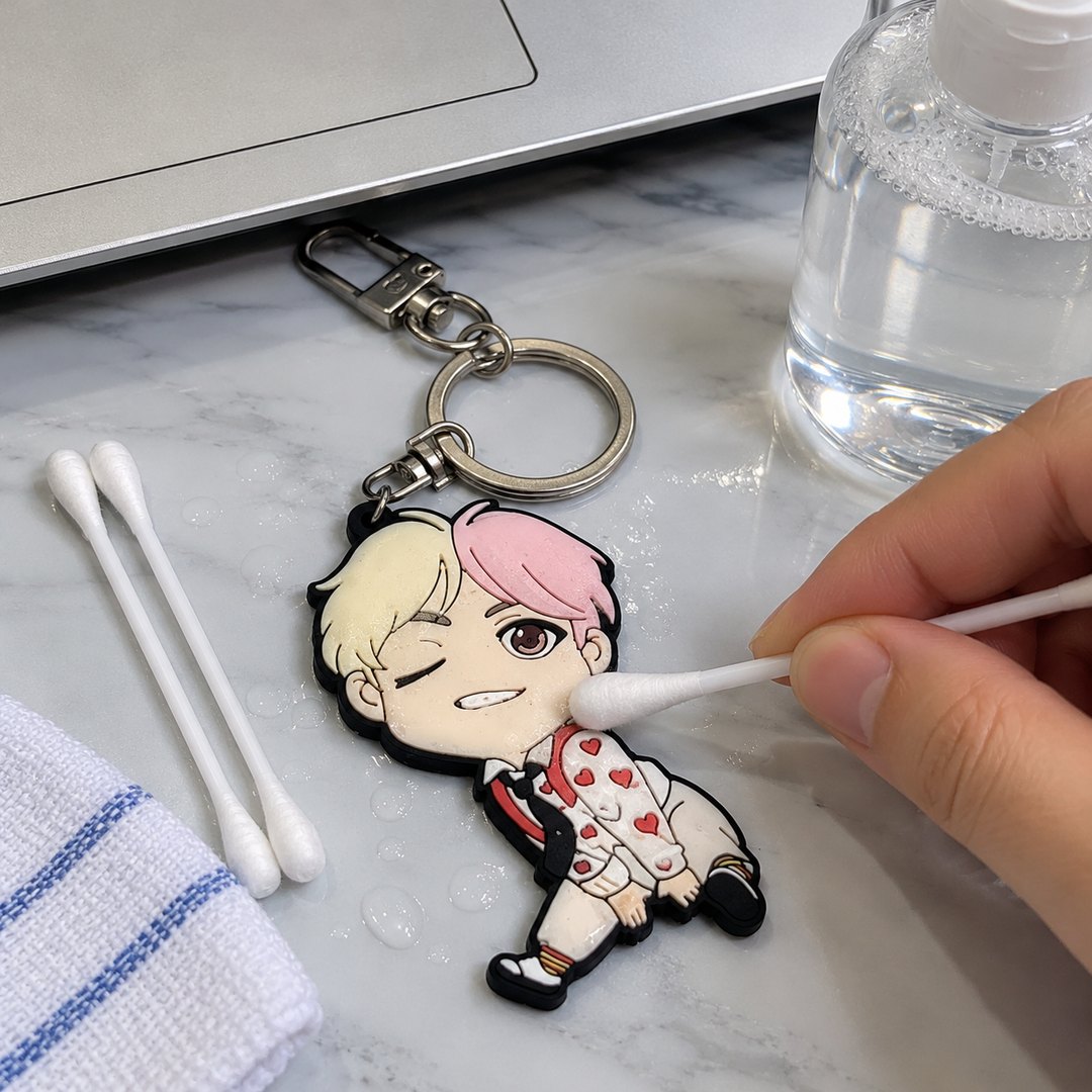 A cotton swab cleaning a soft PVC figure keychain with a pink-haired character design, next to a towel and a bottle of soapy water.