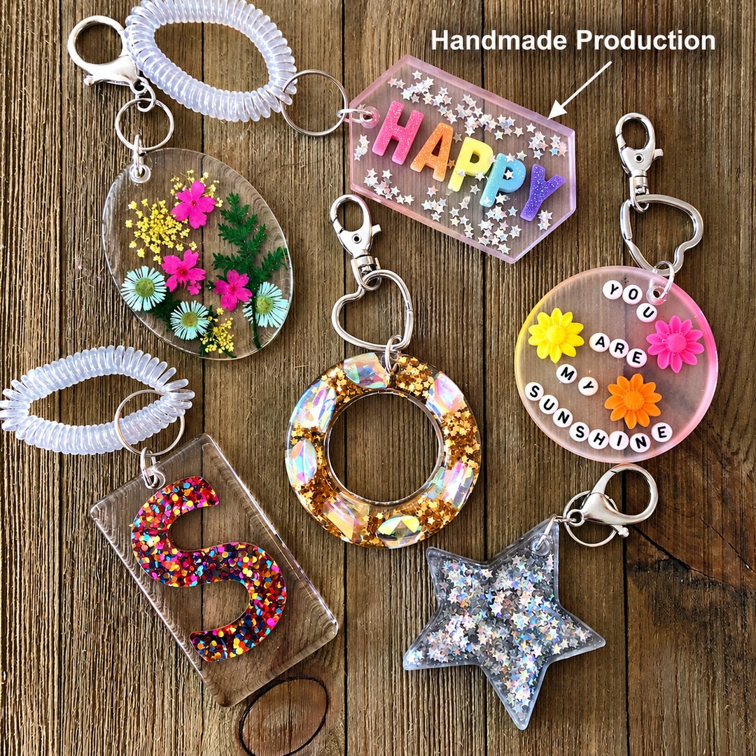 Six handmade resin keychains laid out on a wooden surface, including one reading HAPPY, one reading YOU ARE MY SUNSHINE, a glitter star and pressed flowers.