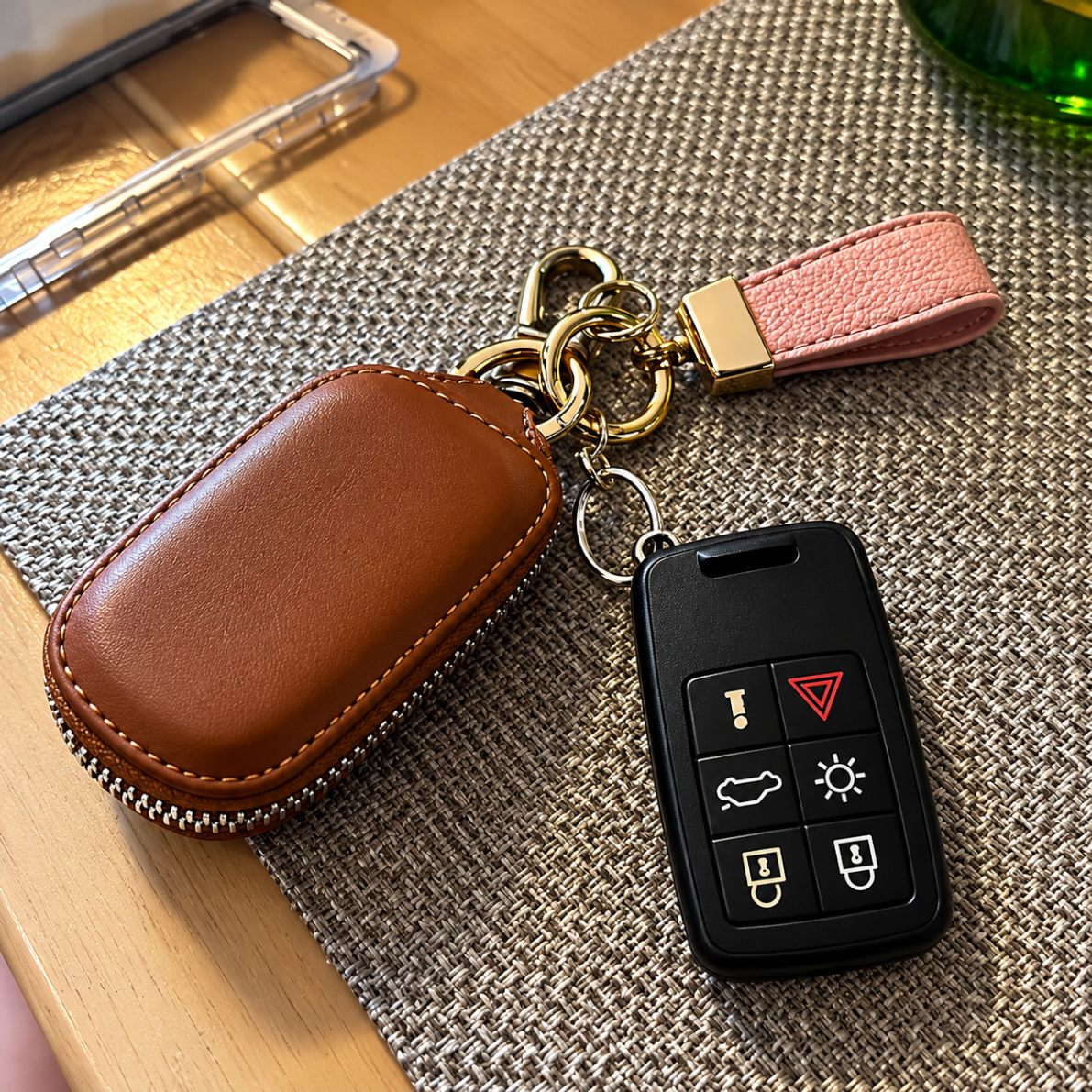 A brown leather car key fob cover beside a black smart key remote and a pink leather strap with a gold clasp on a woven mat