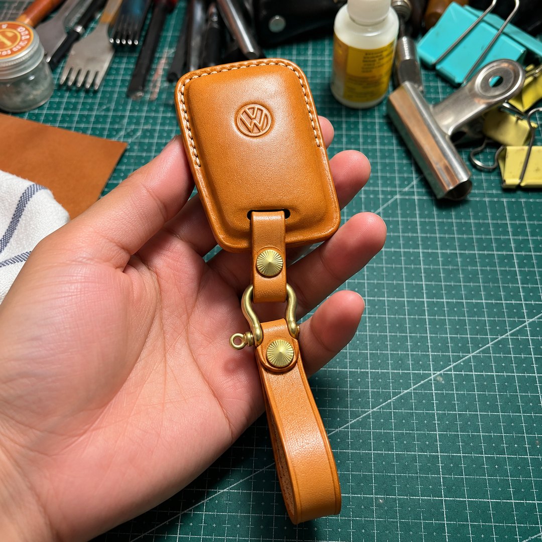 A tan leather key fob with a debossed emblem and brass snap hardware, held in a hand above a leatherwork bench.