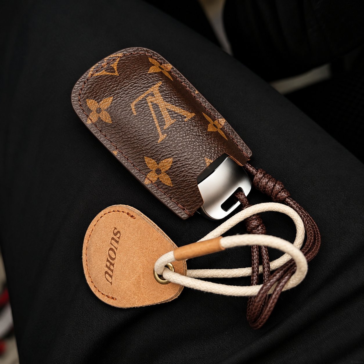 A Louis Vuitton monogram leather car key fob holder with a leather tag stamped SIONS and cream and brown leather cords on a dark seat